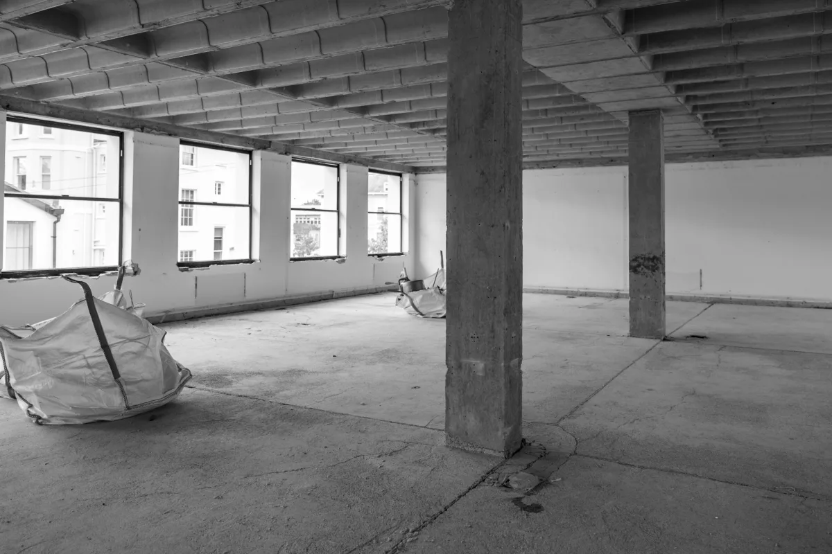 Black and white photograph of a stripped-out floor of a building, with bare concrete columns, a ribbed concrete ceiling, rubble bags on the floor and tall windows looking out onto white town houses